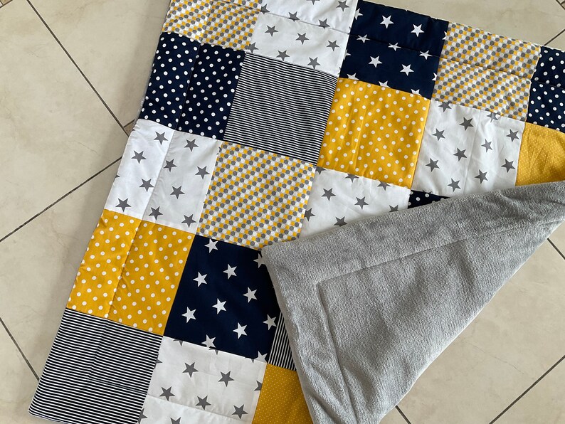 May include: A patchwork baby blanket with a grey fleece backing. The blanket is made of various fabrics in yellow, white, navy blue, and grey with different patterns including polka dots, stripes, and stars.