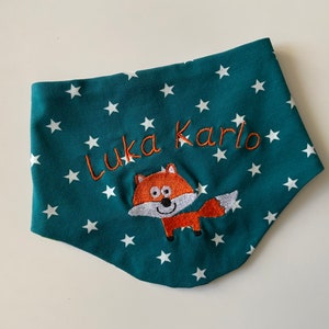 Könnte beinhalten: Ein blaugrüner Stofflätzchen mit weißen Sternen und einem gestickten orangefarbenen Fuchs mit dem Namen "Luka Karlo" in orangefarbenem Garn.