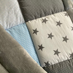 May include: A patchwork baby quilt with a gray and blue color scheme. The quilt is made of different fabrics with various patterns, including polka dots and stars. The quilt is folded in half and has a soft, plush texture.