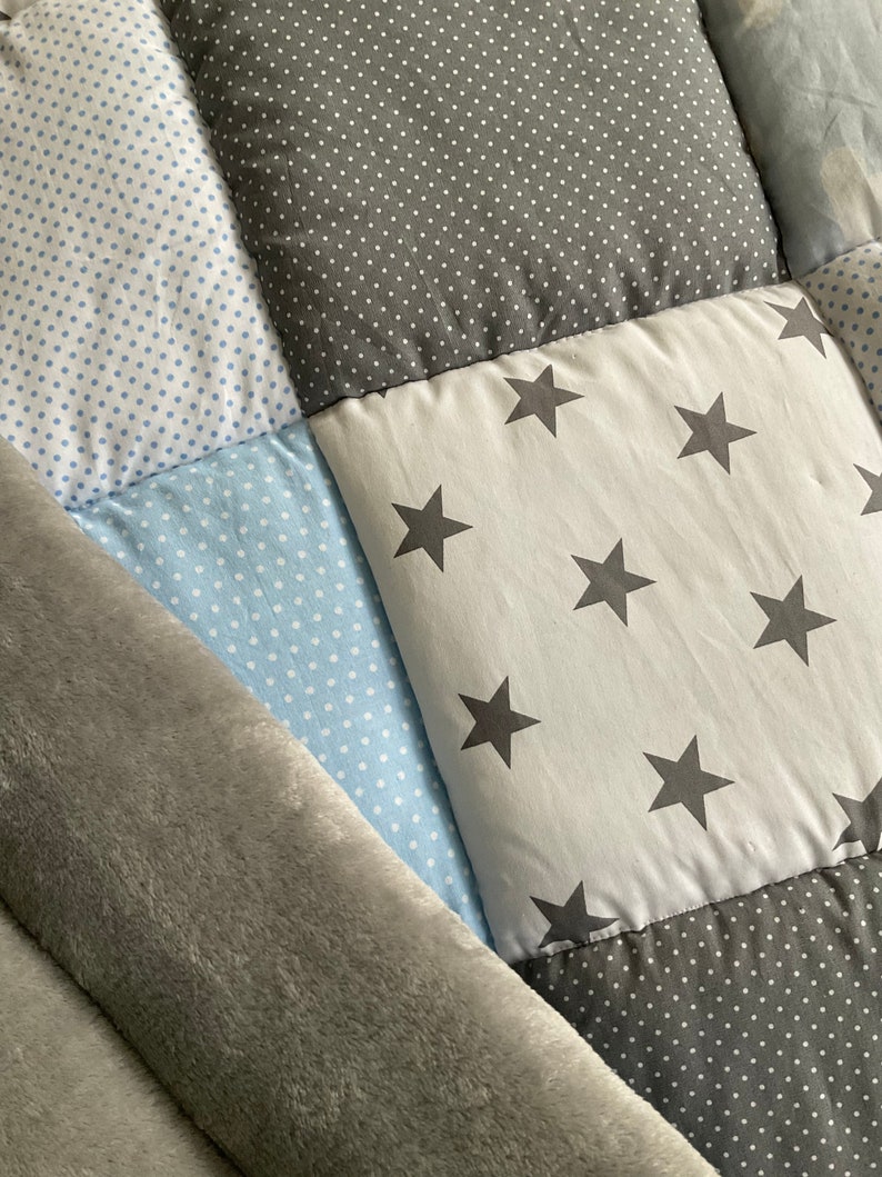 May include: A patchwork baby quilt with a gray and blue color scheme. The quilt is made of different fabrics with various patterns, including polka dots and stars. The quilt is folded in half and has a soft, plush texture.