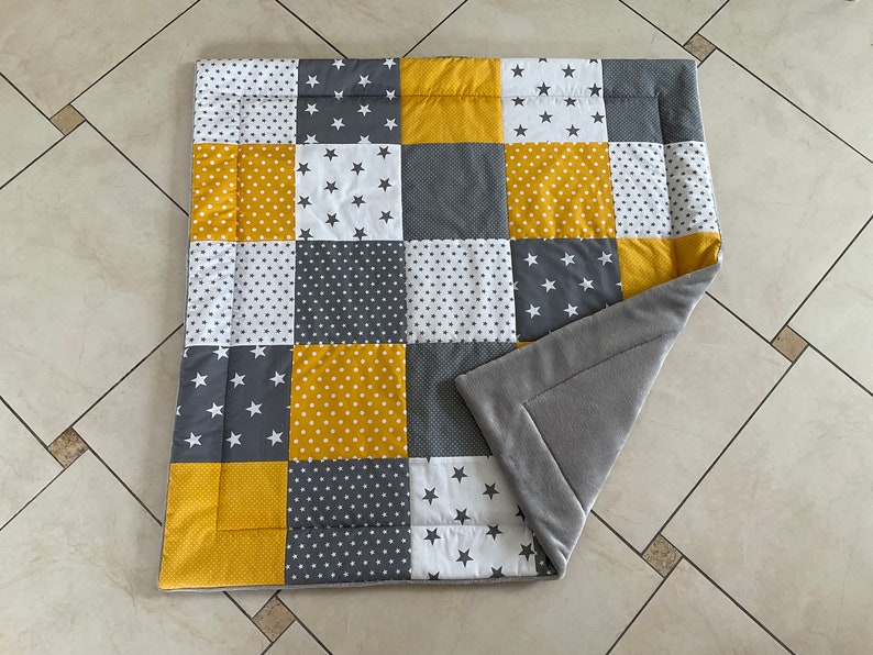 May include: A grey and yellow patchwork baby play mat with a soft grey fleece backing. The mat is made of different fabrics with various patterns, including stars, polka dots, and solid colours.