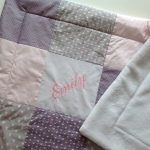 Pu&ograve; includere: Una coperta patchwork per bambini con quadrati di tessuti rosa, grigi e viola. La coperta ha un retro in pile bianco e il nome "Emily" ricamato con filo rosa.