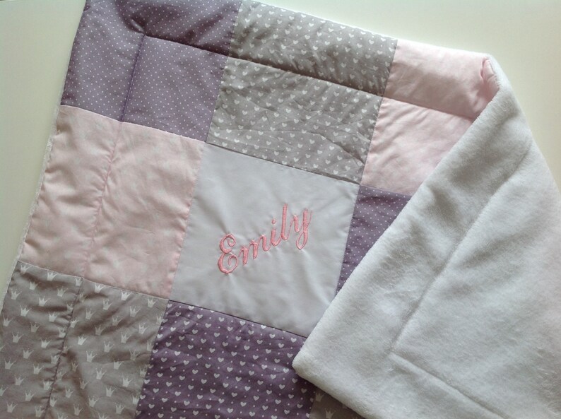 Pu&ograve; includere: Una coperta patchwork per bambini con quadrati di tessuti rosa, grigi e viola. La coperta ha un retro in pile bianco e il nome "Emily" ricamato con filo rosa.