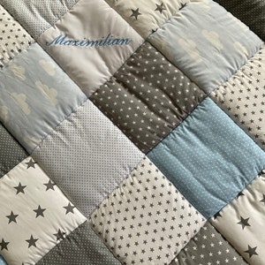 May include: A patchwork baby quilt with a variety of blue and gray fabrics featuring stars and clouds. The quilt is personalized with the name "Maximilian" embroidered in blue thread.
