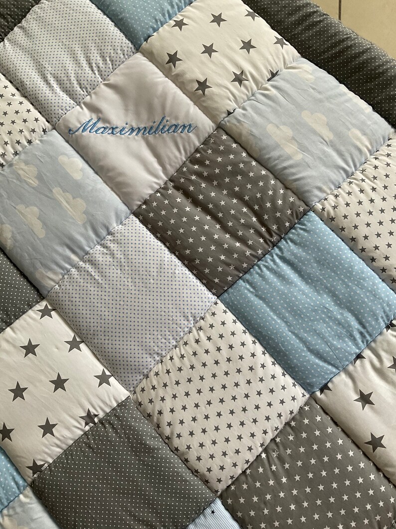 May include: A patchwork baby quilt with a variety of blue and gray fabrics featuring stars and clouds. The quilt is personalized with the name "Maximilian" embroidered in blue thread.