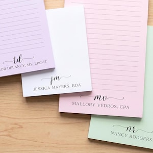 May include: Four rectangular paper notepads in pastel lavender, white, light pink, and mint green. Each notepad features horizontal lines and a personalized monogram with a name and professional title printed in black ink at the bottom.