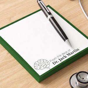 May include: A square notepad with a dark green border and white paper featuring a black line-art brain icon and the printed text "From the desk of Dr. Jack Martin." A black ballpoint pen rests diagonally across the paper, and a stethoscope is visible.