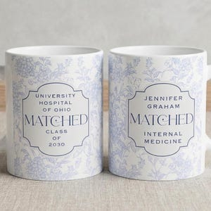 May include: Two white ceramic mugs with a blue floral pattern. Each mug has a label with text: "UNIVERSITY HOSPITAL OF OHIO MATCHED CLASS OF 2030" and "JENNIFER GRAHAM MATCHED INTERNAL MEDICINE".