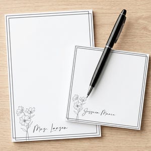 May include: A rectangular notepad and a square notepad, both featuring a black double-line border and a black line-art floral illustration. The rectangular notepad displays the name Mrs. Larsen, while the square notepad displays the name Jessica Marie. A black ballpoint pen rests diagonally across the square notepad.
