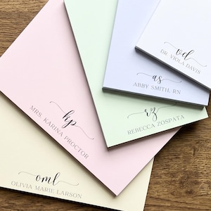 May include: A stack of five rectangular paper notepads in pastel shades of cream, pink, light green, and light blue. Each notepad features a personalized monogram in black script above a name printed in a simple, clean, uppercase sans-serif font.