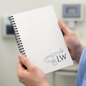 Könnte beinhalten: Ein weißes Spiralnotizbuch mit einer dunkelblauen Stethoskop-Grafik und den Initialen "LW". Das Notizbuch ist geöffnet und zeigt leere Seiten. Der Hintergrund ist eine verschwommene medizinische Umgebung.