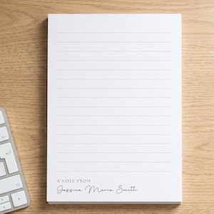 May include: A rectangular white notepad featuring horizontal lines for writing. The bottom section displays the printed text A NOTE FROM followed by the name Jessica Marie Smith in a black script font. The notepad is positioned vertically.
