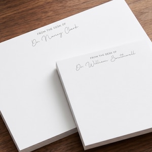 May include: Two stacks of white paper notepads with black printed text. The larger pad reads FROM THE DESK OF Dr. Nancy Clark in a combination of block and script fonts. The smaller pad reads FROM THE DESK OF Dr. William Southwell in the same style.