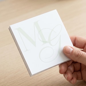 May include: A square stack of white paper sheets featuring a large, light gray monogram with the letters M and G printed in a combination of a bold serif font and a delicate, flowing script font on the top surface of the paper stack.