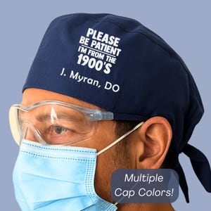 May include: Navy blue surgical scrub cap with the text "PLEASE BE PATIENT I'M FROM THE 1900'S" and "J. Myran, DO" in white. The cap is worn by a person wearing safety glasses and a blue surgical mask. The image also includes the text "Multiple Cap Colors!"