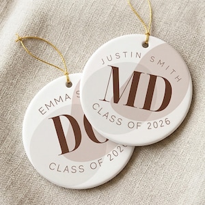 May include: Two round white ceramic ornaments with gold hanging strings. Each features a brown serif monogram, the name Justin Smith or Emma S, and the text Class of 2026, set against abstract beige and light brown organic shapes on the surface.