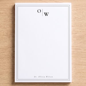 May include: A rectangular white notepad featuring a double-line black border. The top center displays the monogram O | W in a bold serif font. The bottom center features the text Dr. Olivia Wilson in a smaller, elegant serif typeface.