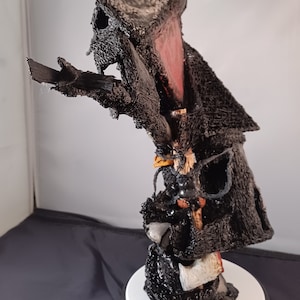Puede incluir: Una escultura negra y gris de una figura con un sombrero grande y un abrigo largo. La figura sostiene un hacha grande en su mano derecha. La escultura está hecha de una variedad de materiales, incluyendo madera, metal y tela. Está montada sobre una base blanca.