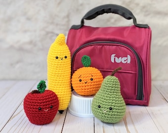 May include: Four crocheted fruit plushies: a red apple, a yellow banana, an orange orange, and a green pear. The plushies have smiling faces and are arranged in front of a pink lunch bag with the word "fuel" on it.