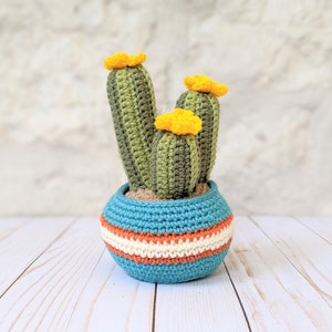 May include: Crocheted cactus in a blue, orange, and white striped pot. The cactus has three green stalks with yellow flowers. The pot is sitting on a white wooden surface.
