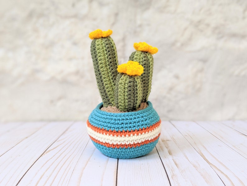May include: Crocheted cactus in a blue, orange, and white striped pot. The cactus has three green stalks with yellow flowers. The pot is sitting on a white wooden surface.