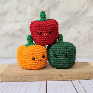 May include: Three crocheted bell peppers, one red, one orange, and one green, are stacked on a wooden cutting board. Each pepper has a cute, embroidered face.