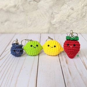 May include: Four crocheted fruit keychains: a blueberry, lime, lemon, and strawberry. Each fruit has a smiling face and a silver keychain ring. The strawberry has a green crocheted stem.