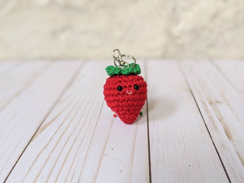 May include: A crocheted red strawberry keychain with a green leafy top and a smiling face. The handmade fruit-shaped charm has a silver-colored clasp for attaching to keys or bags. The background is a light-colored wooden surface.