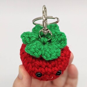 May include: A handmade crochet strawberry keychain. The strawberry is red with green leaves and black eyes. The keychain has a silver clasp.
