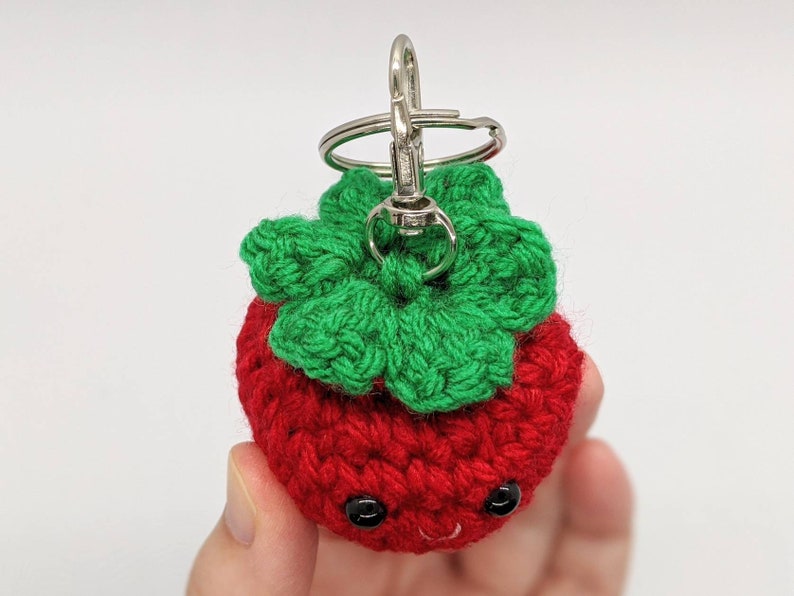 May include: A handmade crochet strawberry keychain. The strawberry is red with green leaves and black eyes. The keychain has a silver clasp.