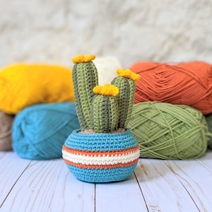 May include: A handmade crochet cactus in a blue pot with orange and white stripes. The cactus is green with yellow flowers. Colorful yarn balls in yellow, teal, orange, and green are in the background.