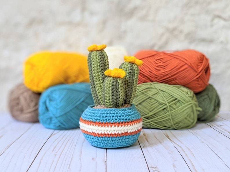 May include: A handmade crochet cactus in a blue pot with orange and white stripes. The cactus is green with yellow flowers. Colorful yarn balls in yellow, teal, orange, and green are in the background.