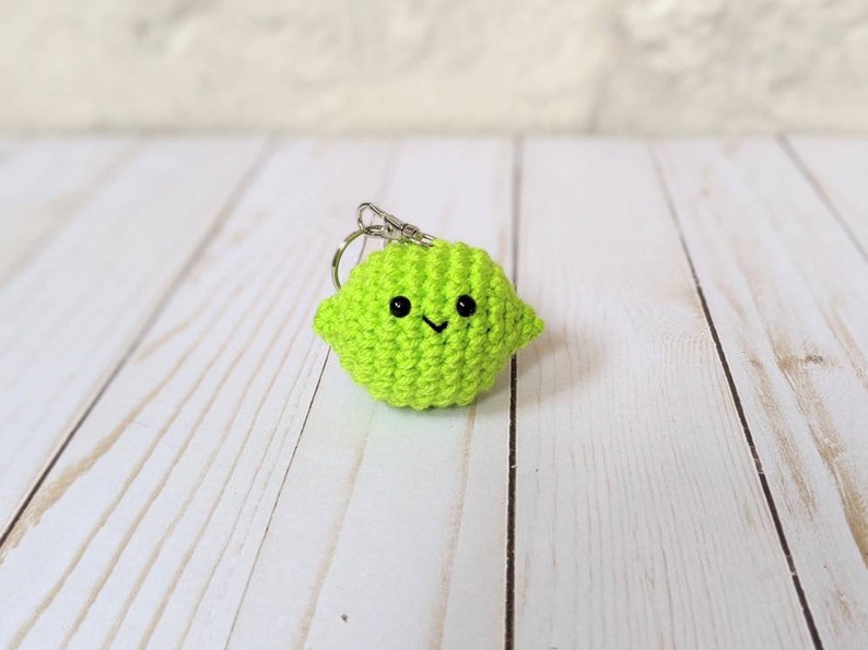 May include: A bright green crocheted lime keychain with a smiling face and a silver-colored keyring. The lime is made of yarn and has a cute, friendly design. The keychain is a fun accessory.