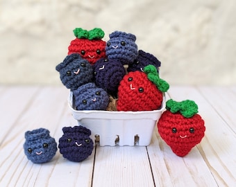May include: A white basket filled with crocheted blueberries and strawberries. The blueberries are blue and purple, and the strawberries are red with green leaves. The crocheted fruits have smiling faces.