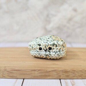May include: A small crocheted stuffed toy shaped like a rock with a light green, beige, and white color scheme. The toy has two black eyes and a small black mouth.