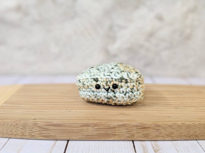May include: A small crocheted stuffed toy shaped like a rock with a light green, beige, and white color scheme. The toy has two black eyes and a small black mouth.