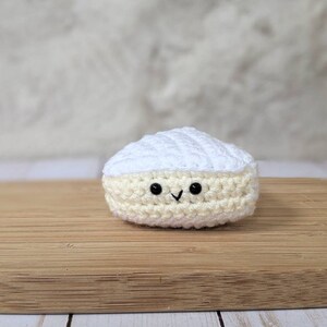 May include: A crocheted white and cream cheese wedge with a smiling face. The cheese is sitting on a wooden cutting board.