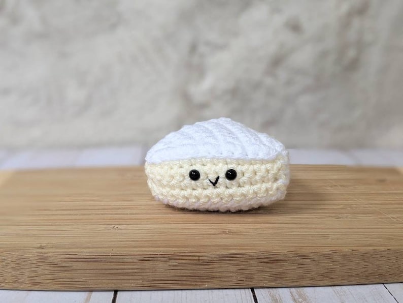 May include: A crocheted white and cream cheese wedge with a smiling face. The cheese is sitting on a wooden cutting board.