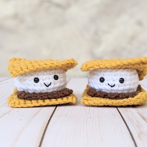 May include: Two crocheted s'mores with smiling faces. The s'mores are made of yellow graham crackers, white marshmallow, and brown chocolate.