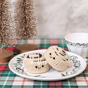 Puede incluir: Dos peluches de rollo de canela tejidos a crochet con caras sonrientes en un plato blanco con hojas verdes y bayas rojas. El plato está sobre un mantel a cuadros rojo y verde.