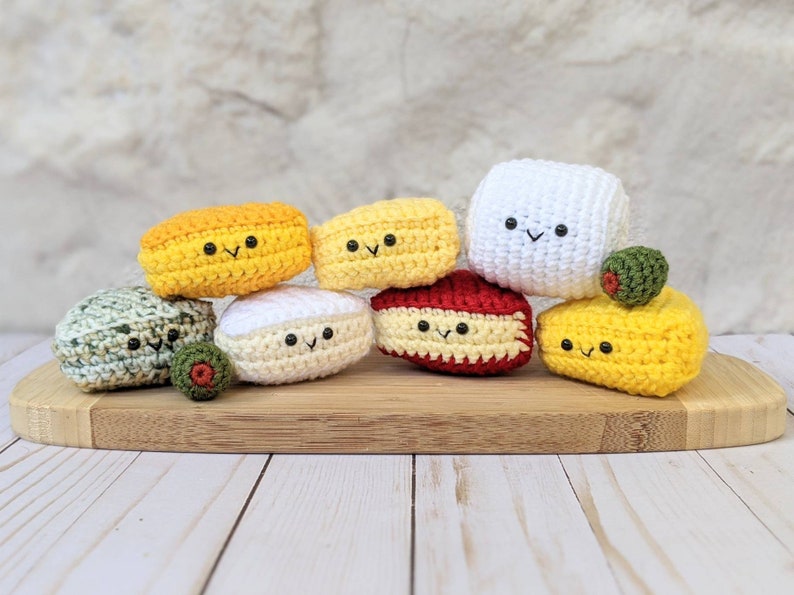 May include: A collection of seven crocheted food items, including a red, white, and yellow cheese, a green olive, and a green and brown cheese. The crocheted food items are arranged on a wooden cutting board.