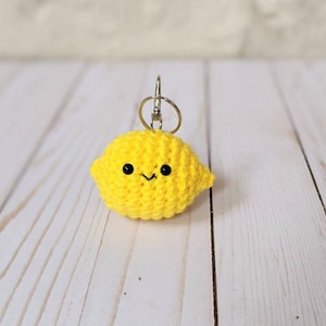 May include: A handmade, yellow crocheted lemon keychain with a smiling face and black eyes. The lemon-shaped charm is attached to a silver-colored keyring. The background is a light-colored wooden surface.
