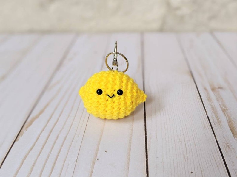 May include: A handmade, yellow crocheted lemon keychain with a smiling face and black eyes. The lemon-shaped charm is attached to a silver-colored keyring. The background is a light-colored wooden surface.
