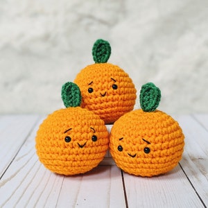 May include: Three handmade, orange crochet pumpkins with green leaf stems. Each pumpkin has a stitched face with a smiling or slightly concerned expression. The pumpkins are arranged on a white wooden surface.