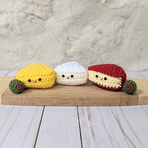 May include: Three crocheted cheese wedges on a wooden cutting board. The cheeses are yellow, white, and red. Each cheese has a smiling face.