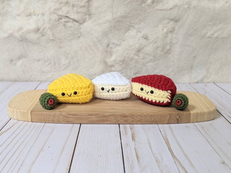 May include: Three crocheted cheese wedges on a wooden cutting board. The cheeses are yellow, white, and red. Each cheese has a smiling face.