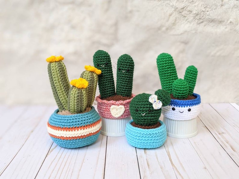 May include: A collection of handmade crochet cactus plants in various sizes and colors. The cacti are in blue, pink, and white pots with smiling faces. Some have yellow flower accents, creating a whimsical and charming display.