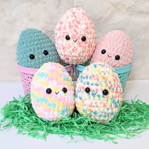 May include: Five crocheted Easter eggs in pastel colors with cute faces. The eggs are in pink and blue baskets and sitting on green grass.