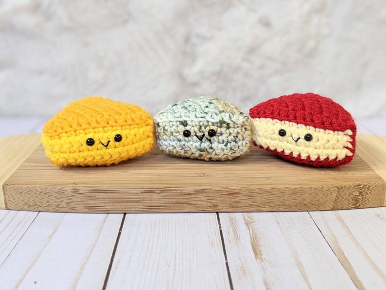 May include: Three crocheted cheese wedges with smiling faces. The cheese wedges are yellow, green and red.