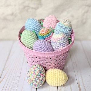 May include: A pink basket filled with colorful, crocheted Easter eggs. The eggs are in various pastel colors, including blue, pink, green, and purple. Two additional eggs sit outside the basket, one speckled and one yellow. The basket is on a white wooden surface.